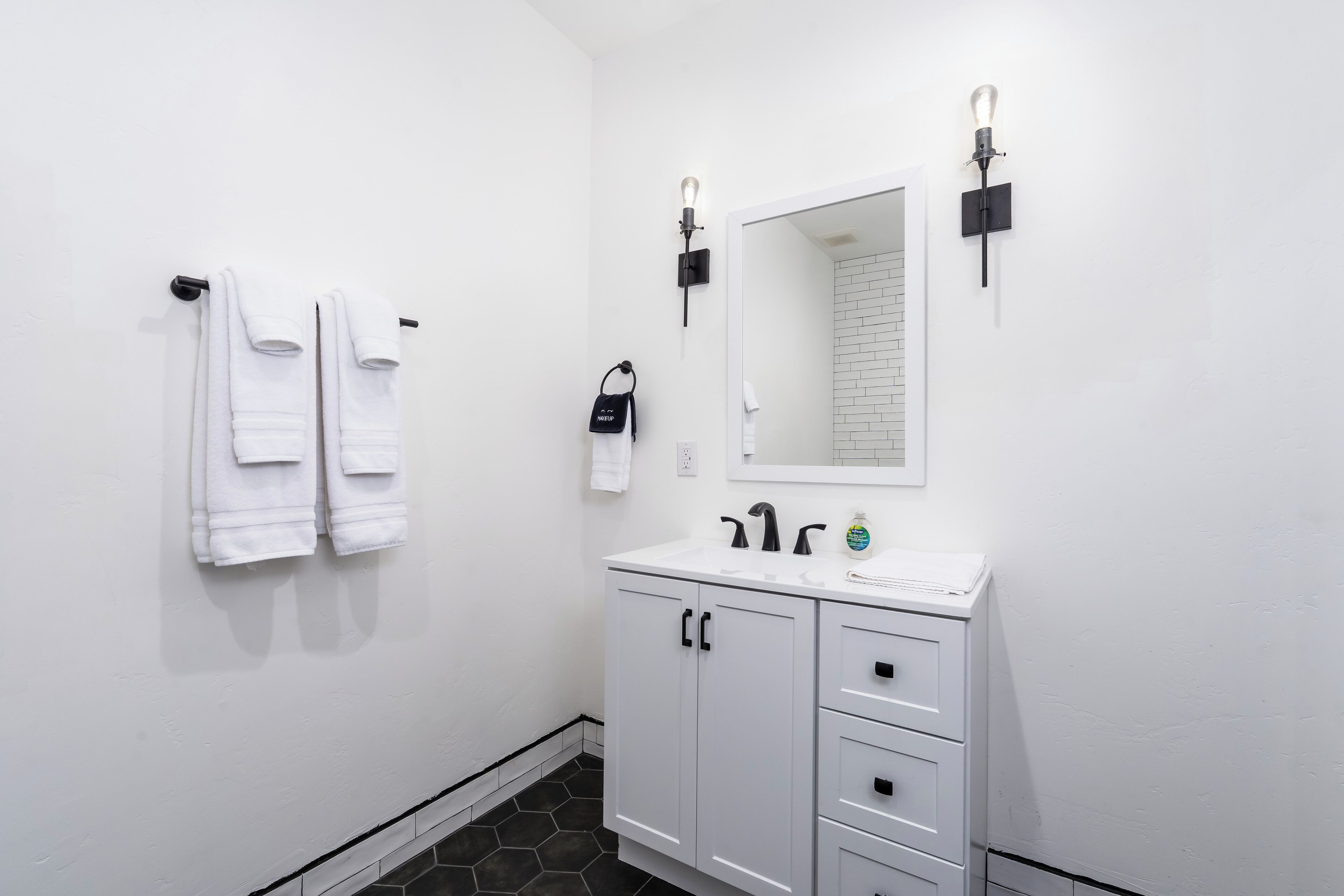 A bathroom with white walls, a white sink, and a mirror above it. There are two towels hanging on a towel rack and a black bag on the wall.