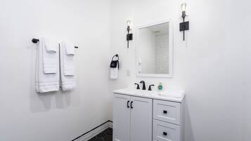A bathroom with white walls, a white sink, and a mirror above it. There are two towels hanging on a towel rack and a black bag on the wall.