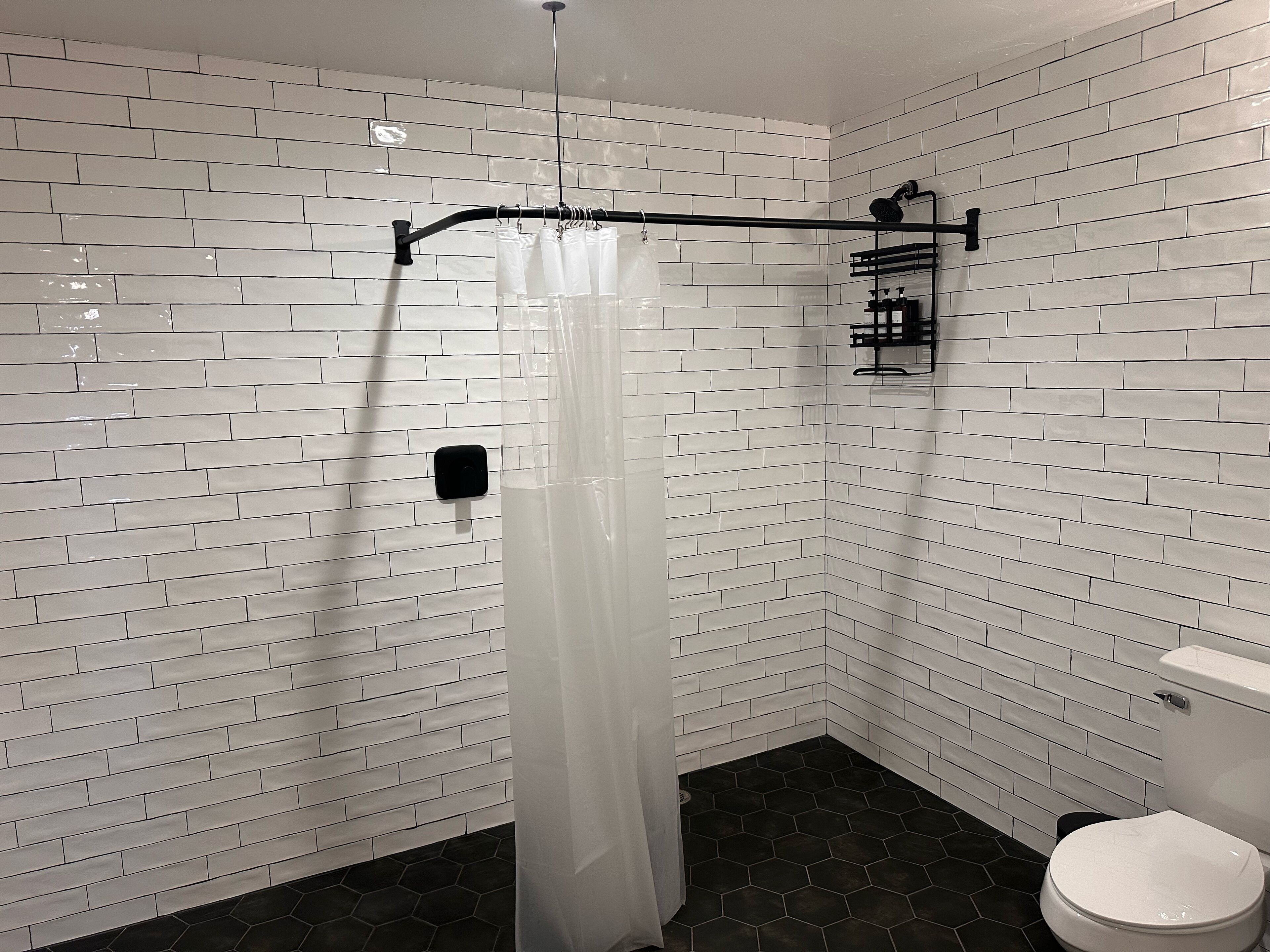 Une salle de bain avec des murs en briques blanches, une douche avec un rideau transparent, un porte-serviettes noir et des toilettes blanches.