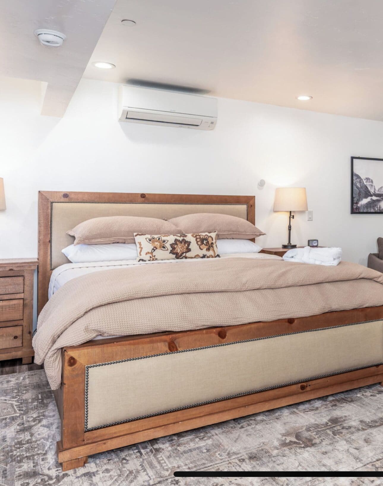 Una cama bien hecha con estructura de madera y ropa de cama color crema, una mesita de noche con una lámpara y un aire acondicionado instalado en la pared.