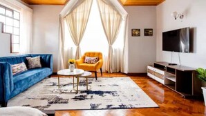 Een woonkamer met een blauwe bank, een gele fauteuil, een salontafel en een tv op een houten dressoir.