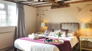 Cama bem arrumada com manta roxa, duas mesas de cabeceira com abajures e uma janela com cortinas.