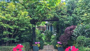 Um caminho de jardim leva a uma casa com cerca de madeira, cercada por vegetação exuberante e vasos com flores vermelhas.