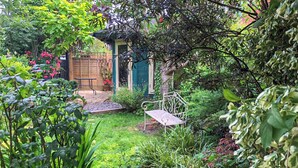 Um jardim exuberante com um espaço de pátio com um banco de metal e uma mesa de madeira.