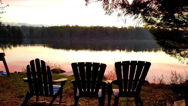 Tres sillas Adirondack mirando un lago tranquilo con una orilla boscosa al fondo.
