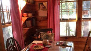 Comedor de una cabaña de madera con una mesa dispuesta para dos personas, sillas, una ventana con cortinas y una estantería con libros y un cuadro enmarcado.
