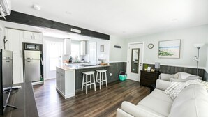 Une cuisine moderne avec des meubles blancs, un îlot central doté de tabourets, un coin repas avec une table blanche et un salon avec un canapé et un téléviseur.