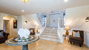 A spacious foyer with a marble staircase, a round table with a decorative centerpiece, and a comfortable seating area with a patterned armchair.
