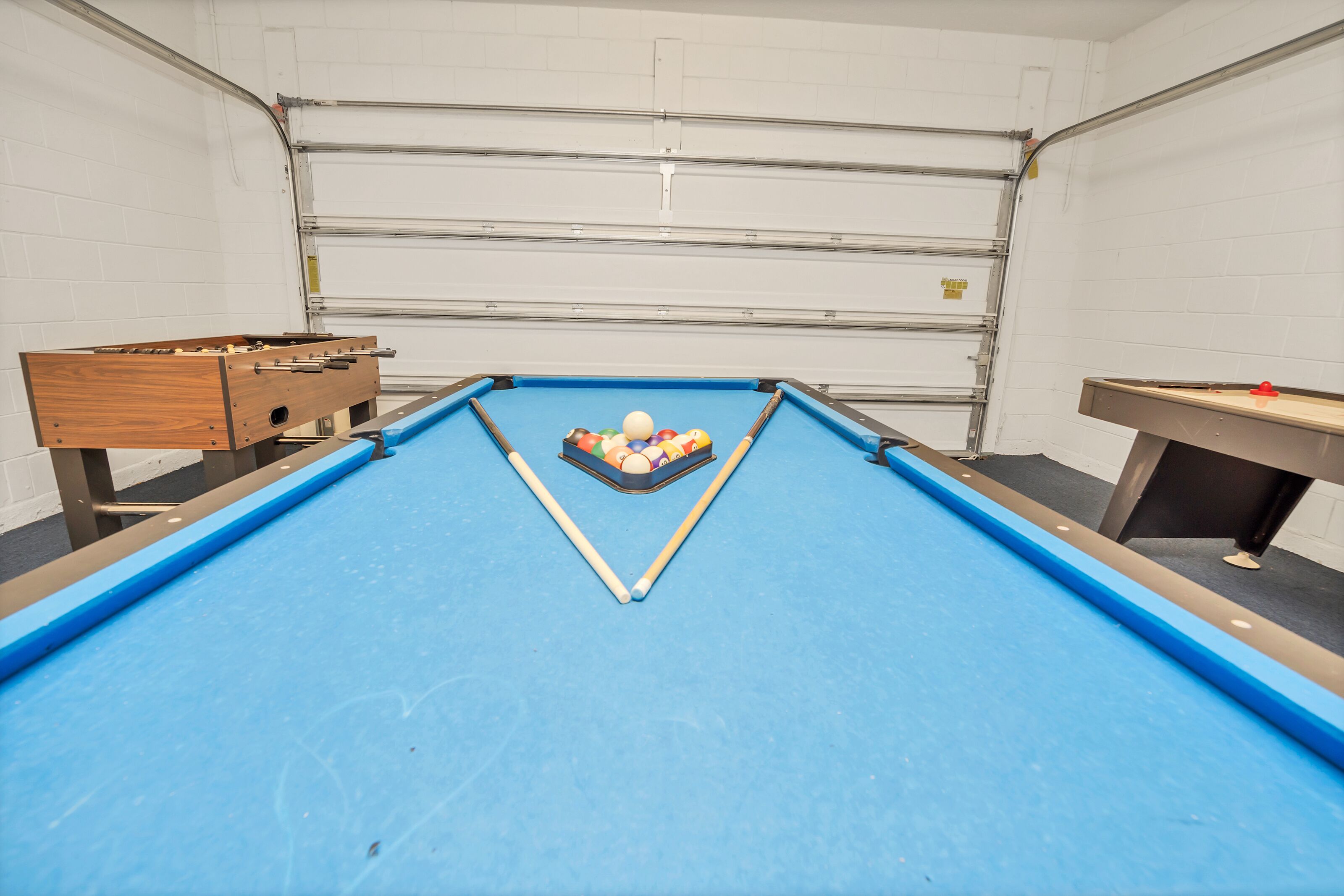 Een pooltafel met ballen, een houten bijzettafeltje en een pingpongtafel in een kamer met witte bakstenen muren en een garagedeur.