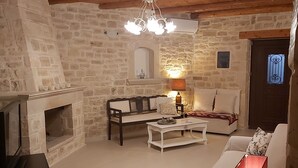 Un salon chaleureux avec une cheminée en pierre, des poutres apparentes en bois au plafond, un canapé blanc, une table basse et un téléviseur posé sur un meuble.