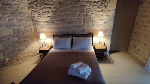 Un lit recouvert d’une couverture marron, deux oreillers blancs et une serviette pliée posée sur le lit. Deux tables de chevet, chacune surmontée d’une lampe, se trouvent de part et d’autre du lit. Les murs de la pièce sont en pierre.