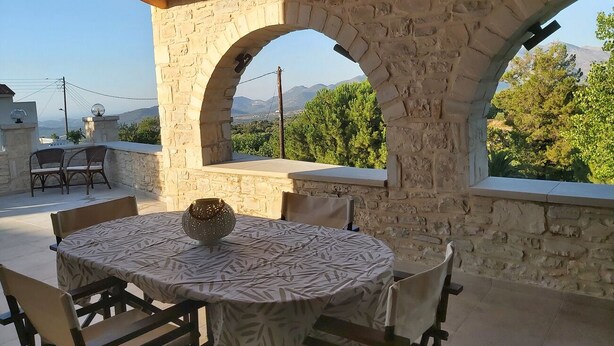 Une terrasse en pierre avec une table ronde et des chaises, un vase décoratif, et une vue sur la verdure et les montagnes, visible à travers des ouvertures en arc de cercle.
