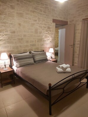 Un dormitorio con pared de piedra, una cama con cabecera, dos mesitas de noche con lámparas y una puerta con cortina.