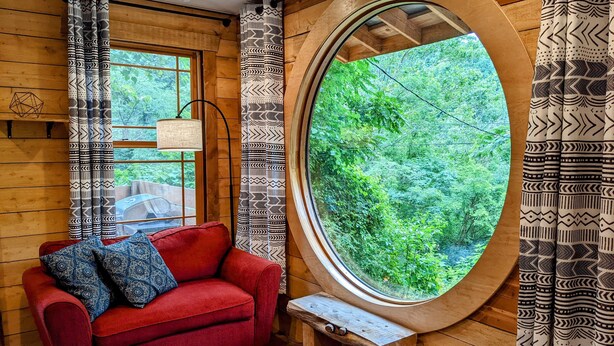 Une pièce chaleureuse avec un canapé rouge, des coussins à motifs, une fenêtre ronde donnant sur un paysage verdoyant et des murs en bois.
