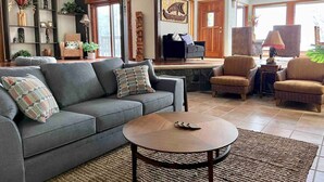 Una sala de estar con un sofá gris, una mesa de centro redonda, sillas de mimbre y una estantería con objetos decorativos.