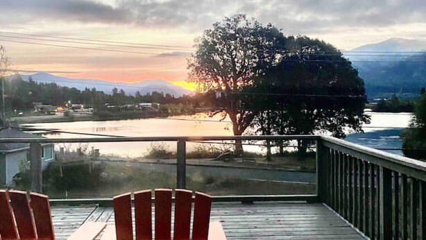 Una terraza de madera con dos sillas Adirondack orientadas a una vista panorámica del atardecer sobre el agua, con montañas al fondo.