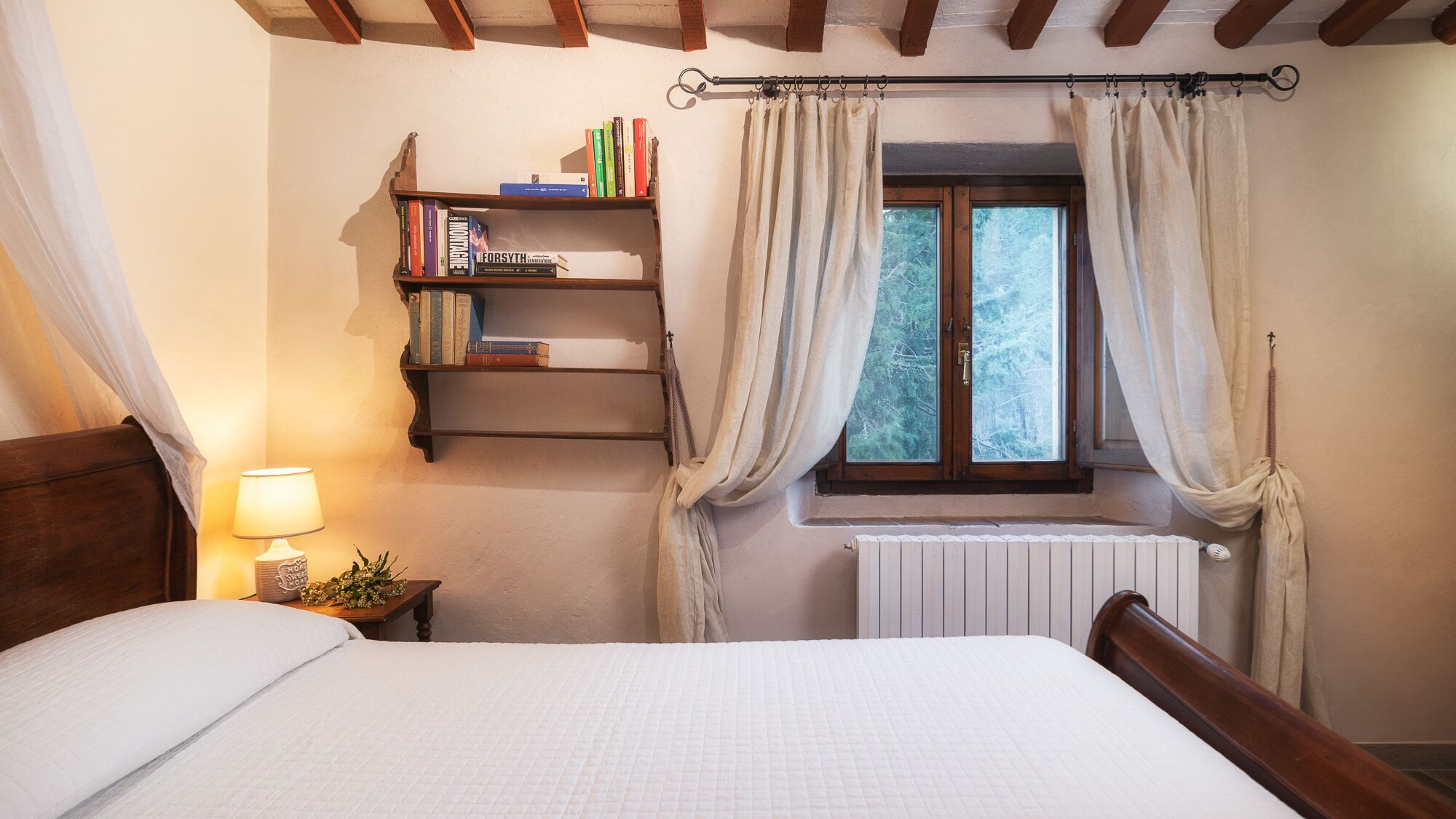 Un dormitorio con cama de madera, una mesita de noche con lámpara, una estantería y una ventana con cortinas.