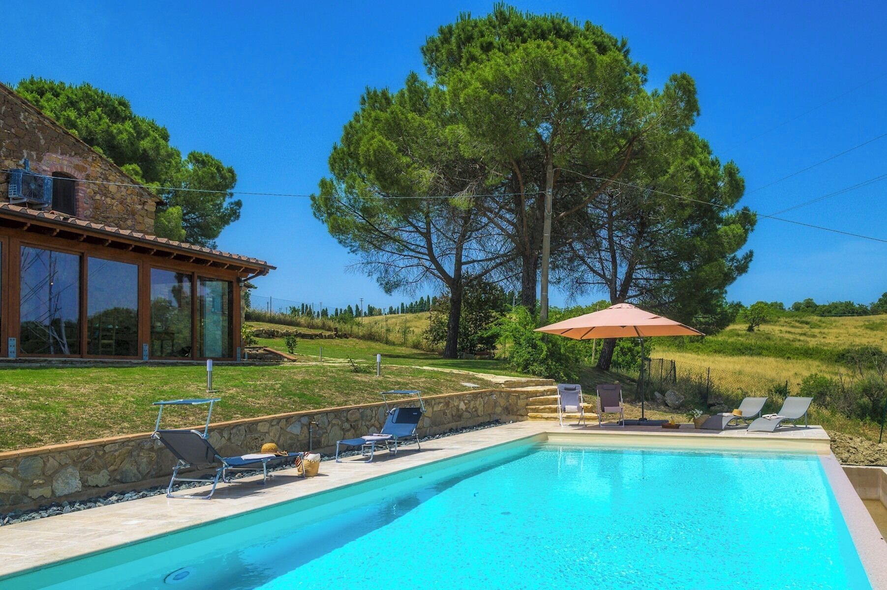 Une piscine entourée d’un mur de pierre et d’un grand arbre, avec des chaises longues et un parasol à proximité.