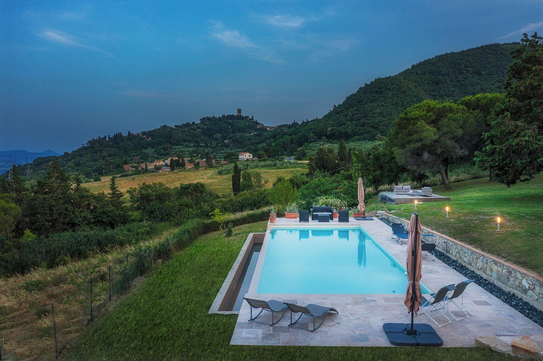A swimming pool with lounge chairs and umbrellas, surrounded by a stone wall and a grassy area, with hills in the background.