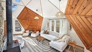 A modern living room with a sloped ceiling, wooden beams, a fireplace, white sofas, and a large window.