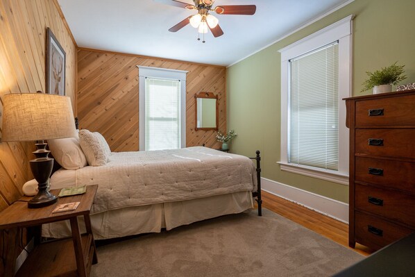 Un dormitorio con cama, paredes de madera, ventilador de techo, cómoda y ventana con persianas.