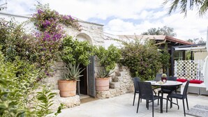 Un patio con mesa y sillas, rodeado de plantas en macetas y un muro de piedra.