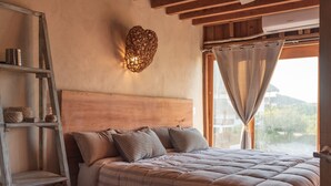 Un dormitorio acogedor con cabecera de madera, cama con ropa de cama a rayas, una lámpara de pared tejida y vistas del exterior a través de una ventana con cortinas.