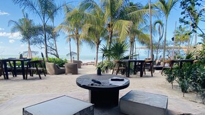 Un área de estar al aire libre con mesas y sillas, rodeada de palmeras y vistas a la playa.