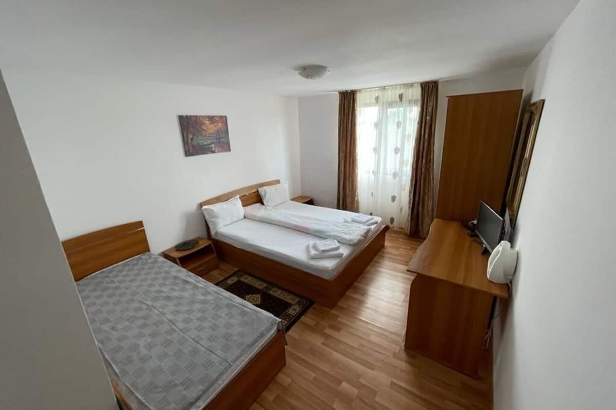 A hotel room with a bed, a desk with a computer, a small table, a chair, and a window with curtains.