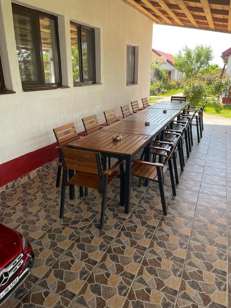 A covered outdoor dining area with a long wooden table and chairs, a tiled floor, and a red car parked nearby.