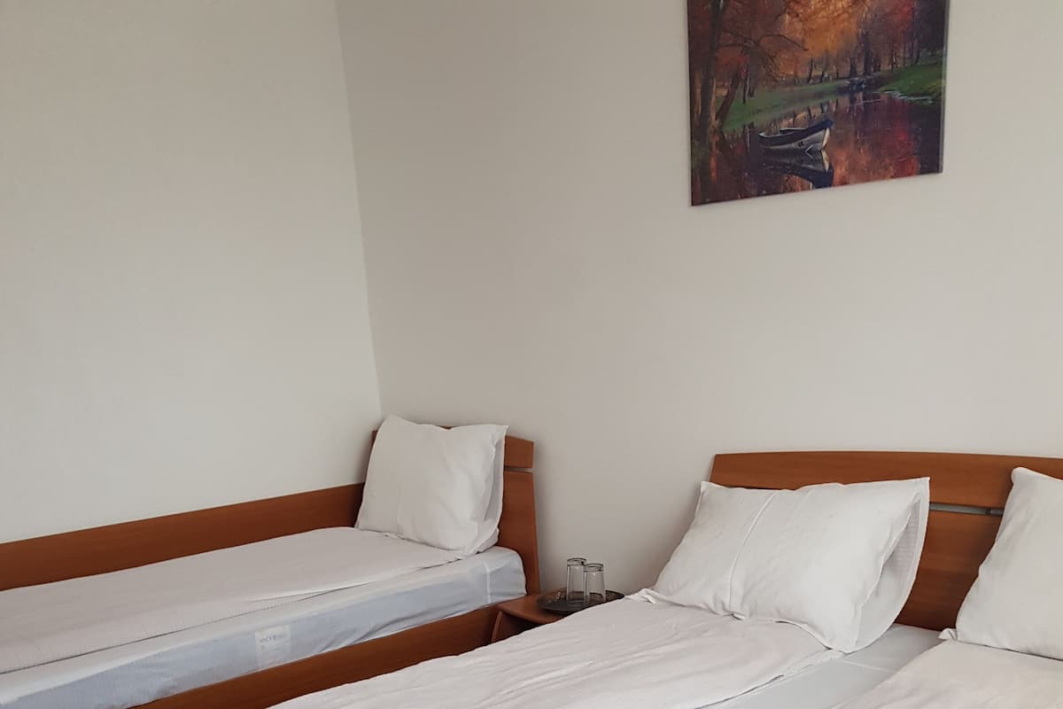 A room with two single beds, wooden headboards, white bedding, a small table with two glasses, and a painting on the wall.