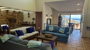 Ampla sala de estar com parede de pedra, um conjunto de sofás, mesa de centro com frutas e vista para o mar através de uma varanda.