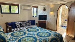 Quarto com cama grande, televisão sobre suporte de madeira, ar condicionado e vista para uma escadaria de pedra.