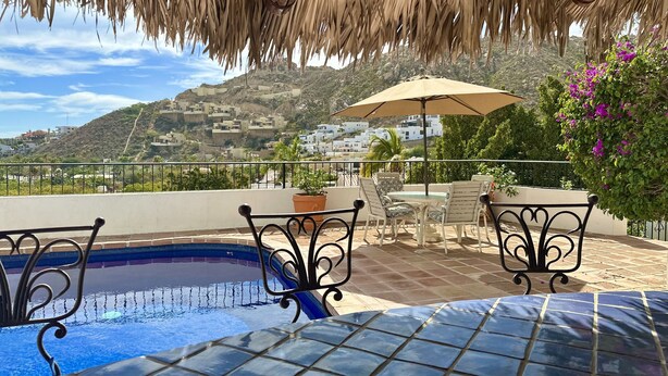 Uma área de piscina com piso ladrilhado, teto de palha e vista para uma encosta com edifícios.