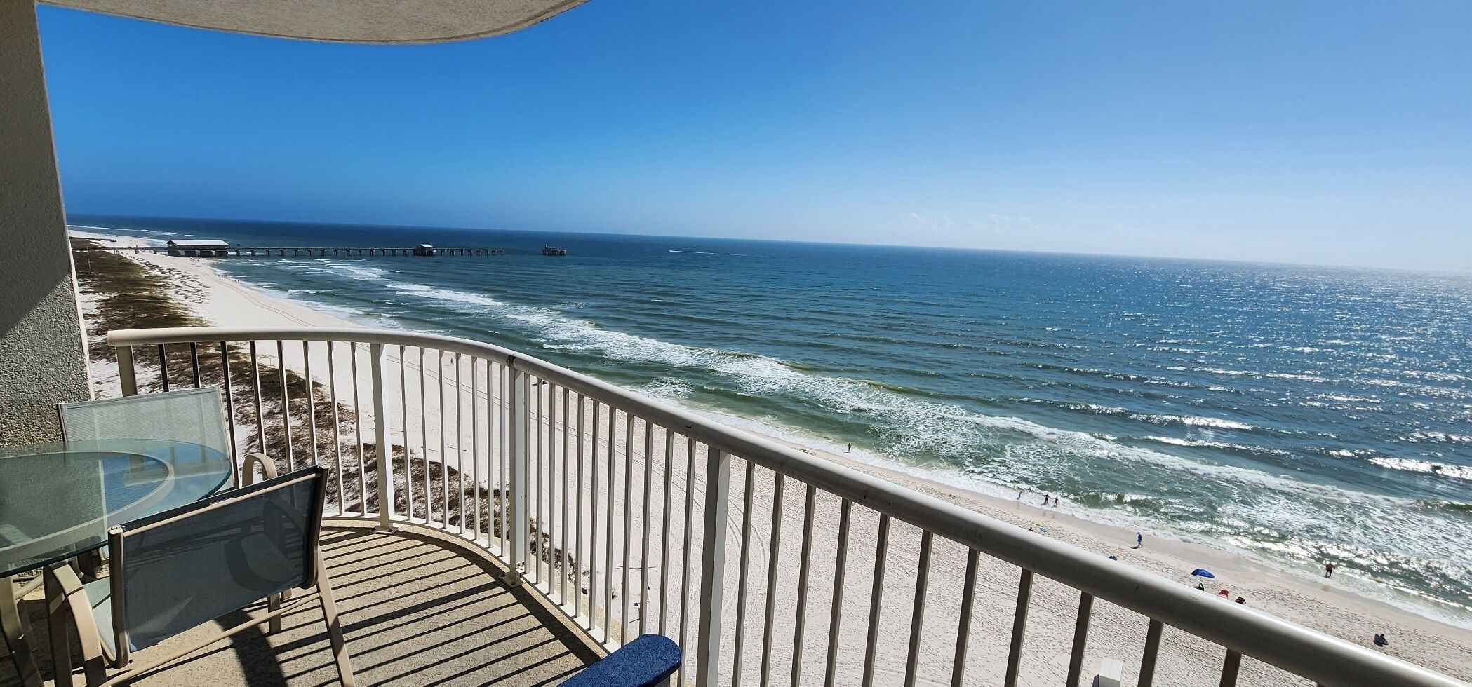 Ein Balkon mit Glastisch und Stühlen, der Blick auf einen Strand mit einem Pier im Hintergrund freigibt.