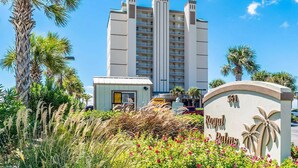 A high-rise hotel with a central tower, surrounded by tropical vegetation and a small building with a sign. - EAST CORNER CONDO DIRECT GULF VIEW/2 BEDROOM & 2 BATH/CLOSE 2 PIER (Gulf Shores)