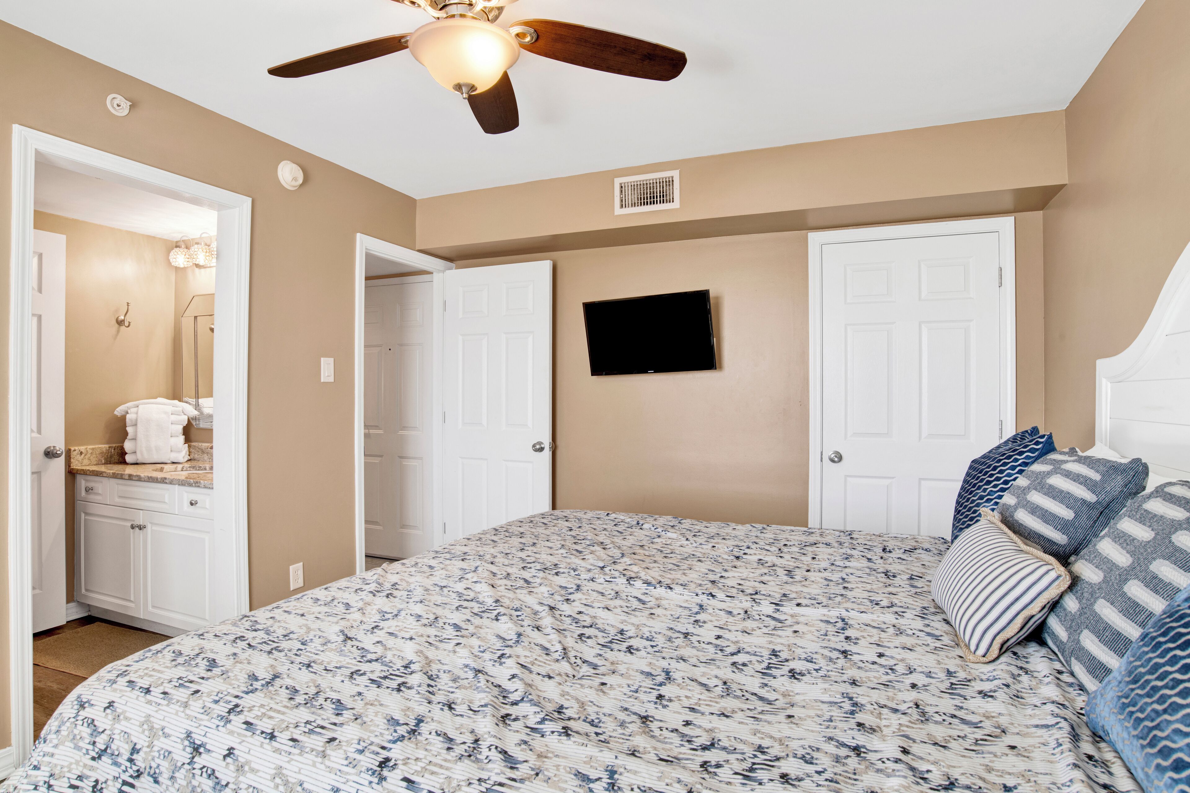 A bedroom with a bed, a ceiling fan, a TV, and a door leading to a bathroom.