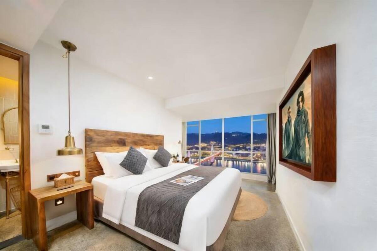 A modern hotel room with a large bed, a wooden nightstand, a bedside table, a large window with a city view, and a framed painting on the wall.