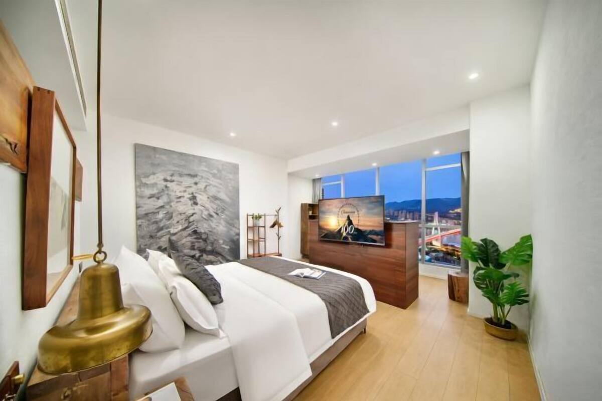 A modern bedroom with a bed, a painting, a TV, and a large window with a city view.