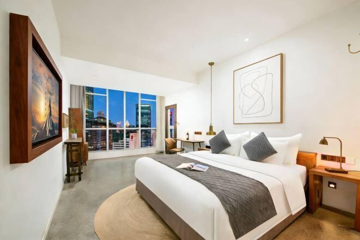 A modern hotel room with a large bed, a desk, a chair, and a view of the cityscape through large windows.