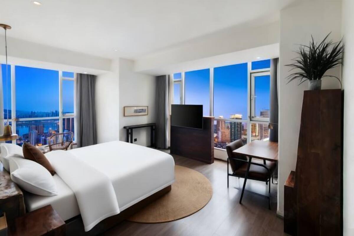A modern hotel room with a large bed, a desk, a chair, a TV, and a view of the cityscape.