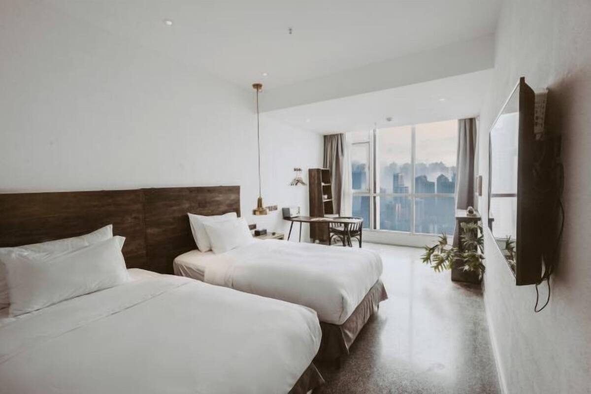A modern hotel room with two beds, a desk, and a large window offering a city view.