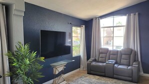 Ein Wohnzimmer mit einem großen Flachbildfernseher an einer strukturierten Wand, einem grauen Sofa mit verschiedenen Sitzgelegenheiten, einem kleinen Beistelltisch, einer Topfpflanze und einem großen Fenster mit Vorhängen.