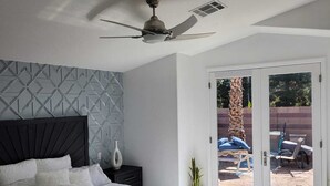 Ein modernes Schlafzimmer mit einem großen Bett, einem Deckenventilator und einer Glastür, die zu einem Außenbereich mit Stühlen und einer Palme führt.