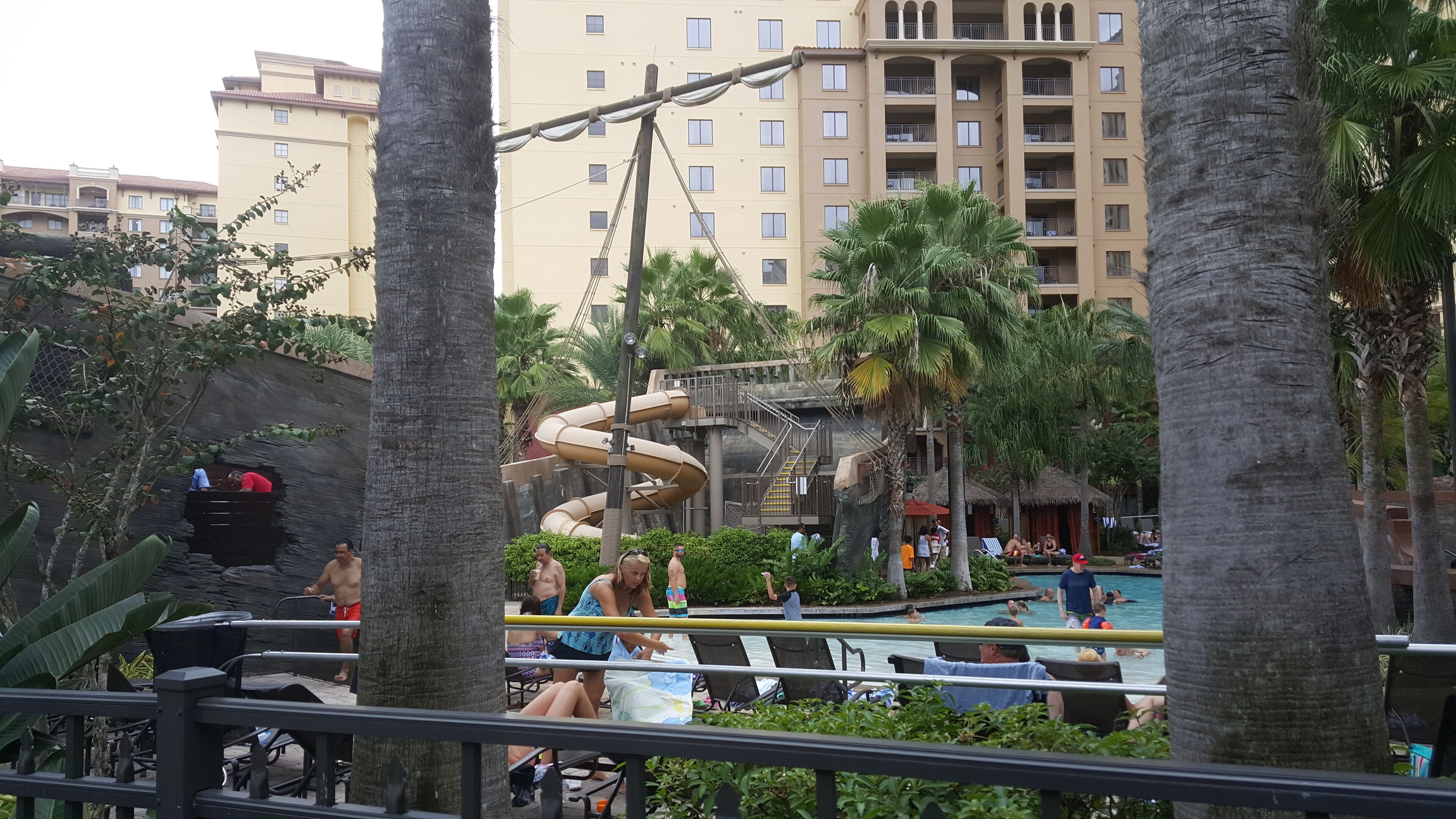 A resort pool area with a water slide, palm trees, and a multi-story building in the background.