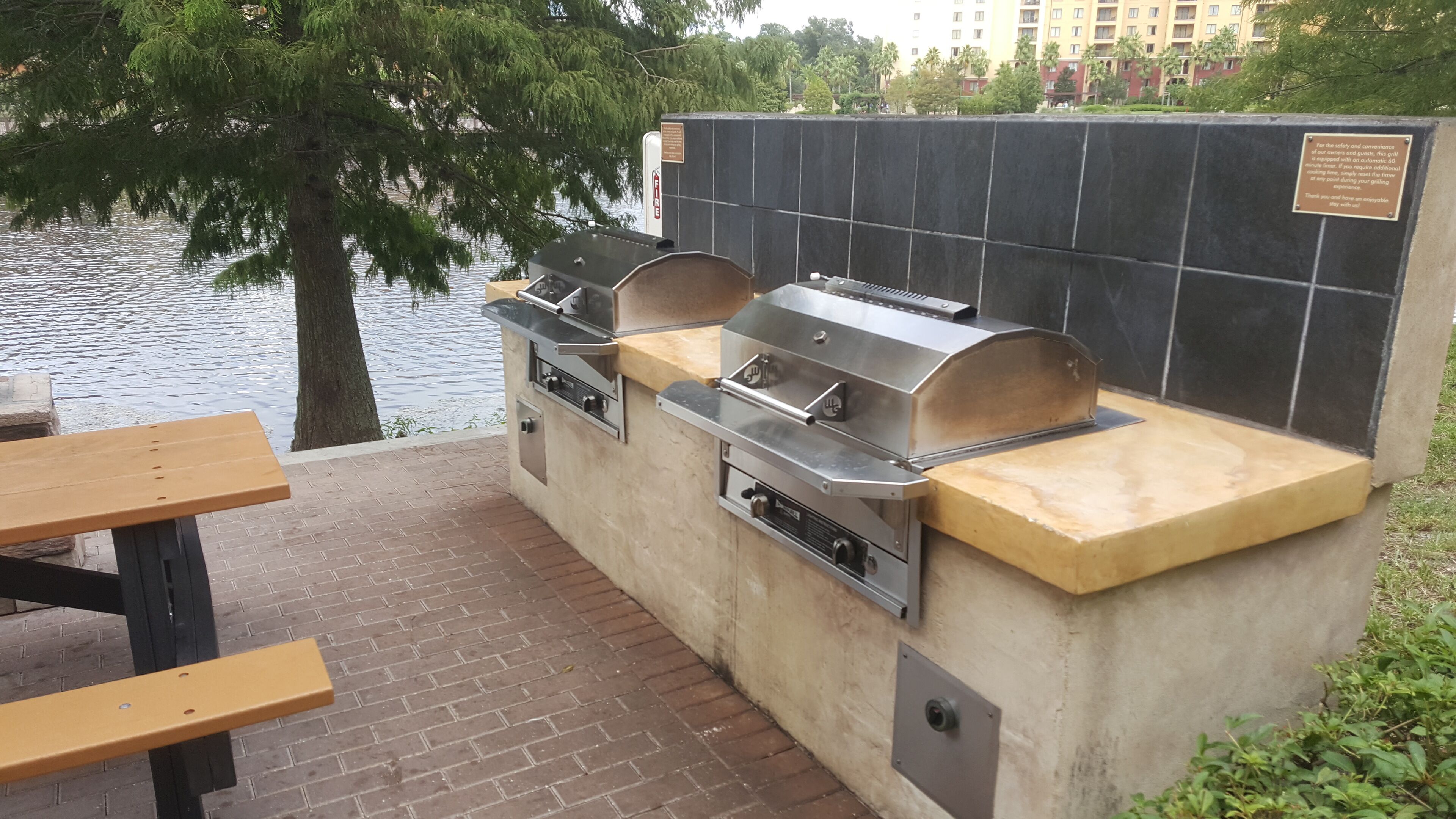 Ein Grillplatz im Freien mit gefliestem Boden und einem Holztisch, in der Nähe von Wasser mit Bäumen im Hintergrund.