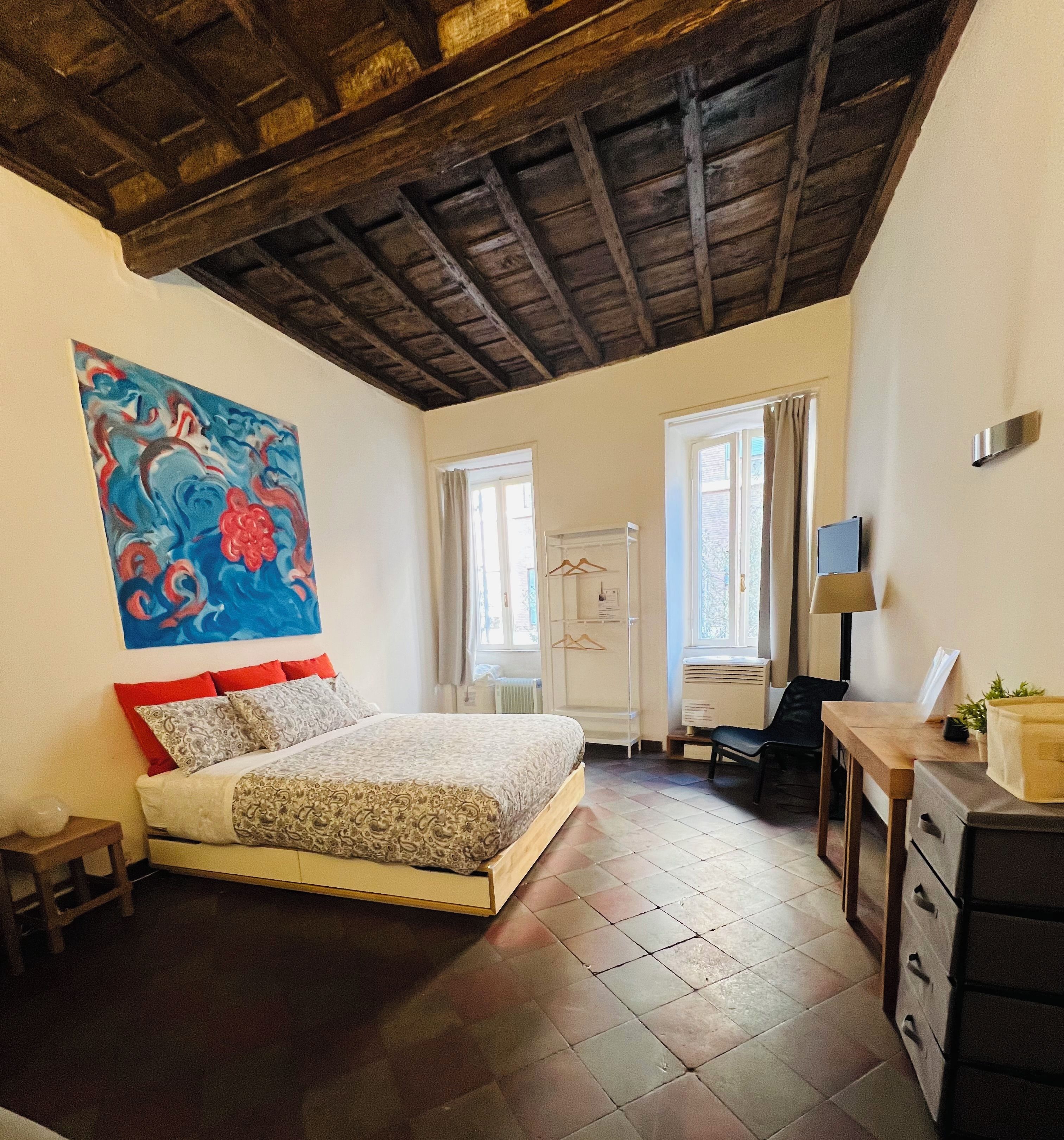 A bedroom with a bed, a painting, a window, a heater, and a desk.