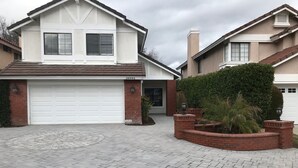 A two-story house with a white garage door, a brick driveway, and a landscaped front yard with a palm tree.