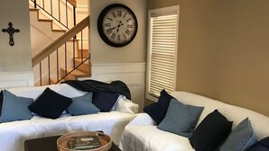 A living room with two white sofas, a black ottoman, a wall clock, and a staircase in the background.