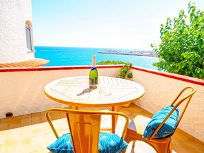Une table ronde en bois, sur laquelle se trouvent une bouteille et un verre, deux chaises jaunes avec des coussins bleus, et une vue sur la mer et une côte lointaine.
